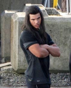 a young man with long hair standing in front of a cement wall and holding his arms crossed