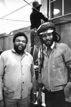 two men standing next to each other in front of a building and one is wearing a helmet