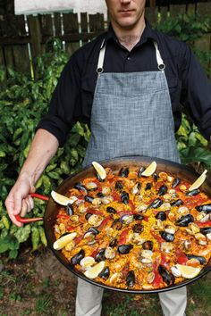 a man in an apron holding a large pan filled with seafood and mussels