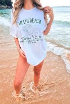 a beautiful woman standing on top of a sandy beach next to the ocean wearing a white shirt