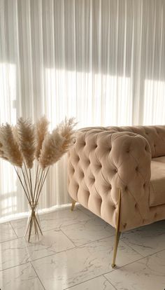 a couch sitting in front of a window next to two tall dry grass stalks on top of a white tiled floor