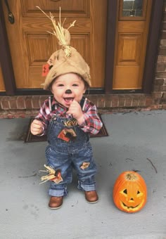 a baby wearing overalls and a hat standing in front of a door next to a pumpkin