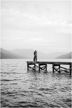 black and white photo of couple kissing on pier over water with mountains in the background