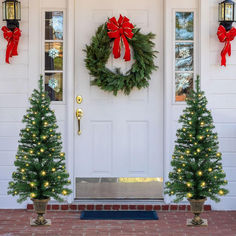 two christmas trees are in front of a white door with red bows and lights on them