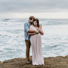 a man and woman embracing each other while standing on top of a cliff near the ocean