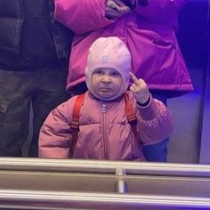 a small child wearing a pink jacket and hat standing in front of a window with other people