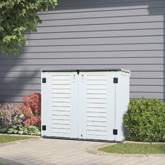 a white storage shed sitting in front of a gray house next to a flower garden