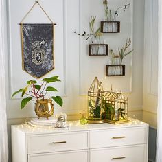 a white dresser topped with lots of drawers and plants in front of a wall hanging