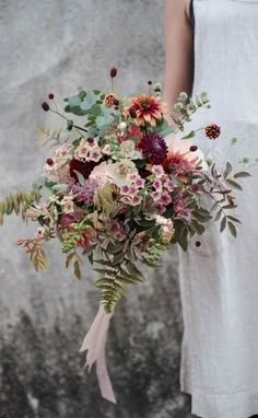 a woman holding a bouquet of flowers in front of a stone wall with leaves and foliage