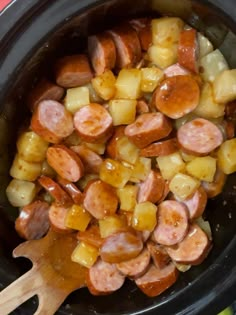 a slow cooker filled with cooked sausage and pineapples next to a wooden spoon