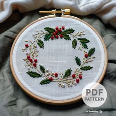 a cross stitch wreath with red berries and green leaves on white fabric in front of a white towel