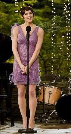 a woman in a purple dress standing next to a microphone on stage with lights behind her