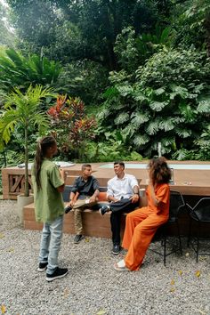 three people sitting on a bench talking to each other in front of some trees and bushes