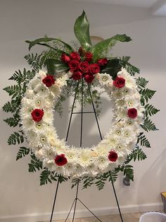 a white wreath with red roses and greenery in the shape of a heart on a tripod stand