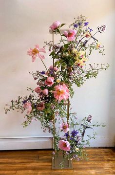 a tall metal vase filled with flowers on top of a hard wood floor next to a white wall