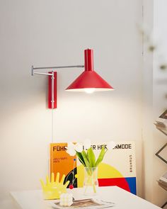 a red lamp on the wall above a white table with a vase and flowers in it