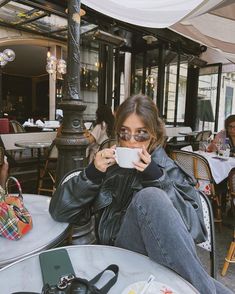 a woman sitting at an outdoor table drinking from a coffee cup while holding a camera