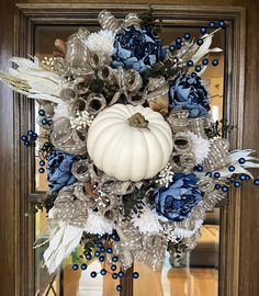 a white pumpkin surrounded by blue flowers and feathers on a front door frame with an autumn wreath