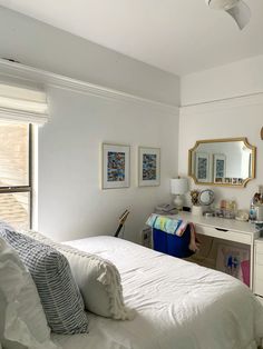 a white bed sitting under a window next to a desk with a mirror on it