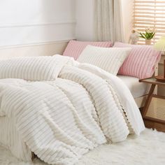 a white bed with pink pillows and blankets on it in front of a windowsill