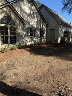 a white house sitting in the middle of a dirt field next to a tree and grass covered yard