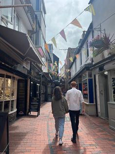 two people walking down an alley way with shops on both sides and flags flying overhead