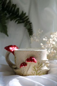 a white cup with red mushrooms on it sitting on a bed next to a plant