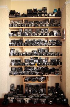 a shelf filled with lots of cameras on top of a wooden floor next to a wall