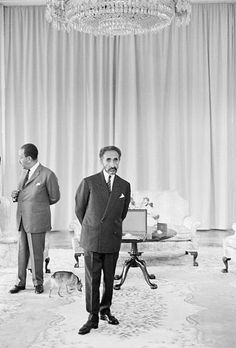 two men in suits standing next to each other near a chandelier and couch