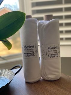 two white towels sitting on top of a wooden table next to a plant and window