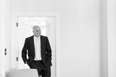 an older man in a suit and tie standing at the door to his room, looking into the hallway