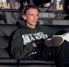 a man sitting on top of a black couch wearing a green hoodie and sneakers