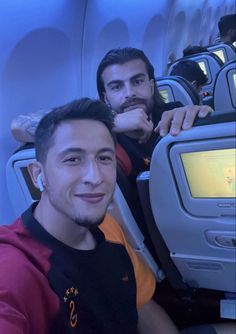 two men are sitting on an airplane and one is looking at the camera while the other man smiles