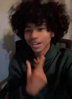 a young man with curly hair sitting in a chair and making the peace sign while wearing a green shirt