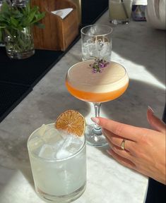 a person holding up a glass with an orange slice in it and another drink on the table