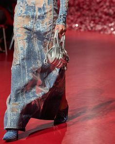 a woman is walking down the runway carrying a handbag in her right hand and wearing blue jeans