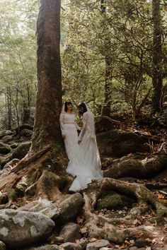 two brides standing next to each other near a tree in the middle of a forest