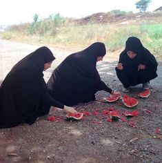 three women in burkas eating watermelon slices on the side of a road