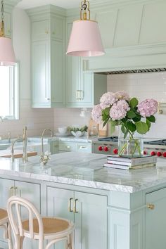 a kitchen with two pink lamps hanging from the ceiling and flowers in a vase on the island