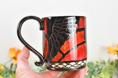 a hand holding a black and red coffee mug with designs on the outside, in front of flowers