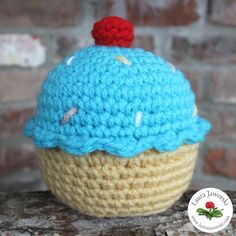 a crocheted cupcake with a red cherry on top sits on a piece of wood