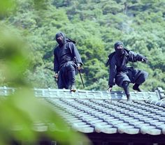 two men in black ninja garb walking on a roof with trees in the background