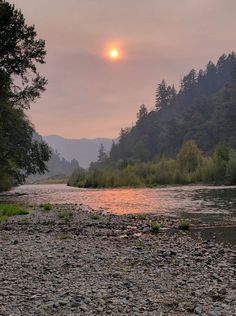 the sun is setting over a river with rocks and trees in the foreground,