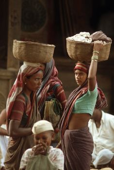 two women carrying baskets on their heads in front of people sitting down and standing around