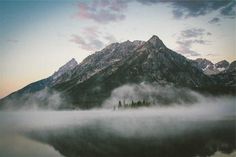 the mountains are covered in clouds and mist as the sun sets over them on a lake