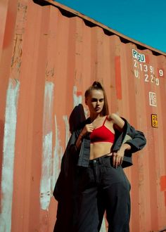 a woman in a red top and black pants standing next to a rusted wall