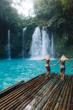 two women standing on bamboo rafts in front of a waterfall with blue water and greenery