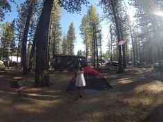 a woman standing in front of a tent next to trees and camping equipment on the ground