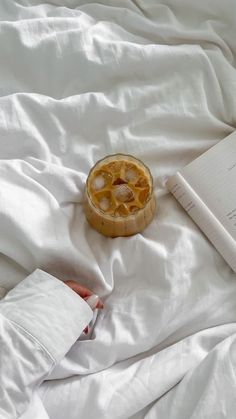 an open book on a bed with white sheets and a fruit tart next to it