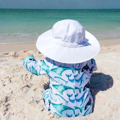 a little boy sitting in the sand at the beach wearing a hat and looking out into the ocean